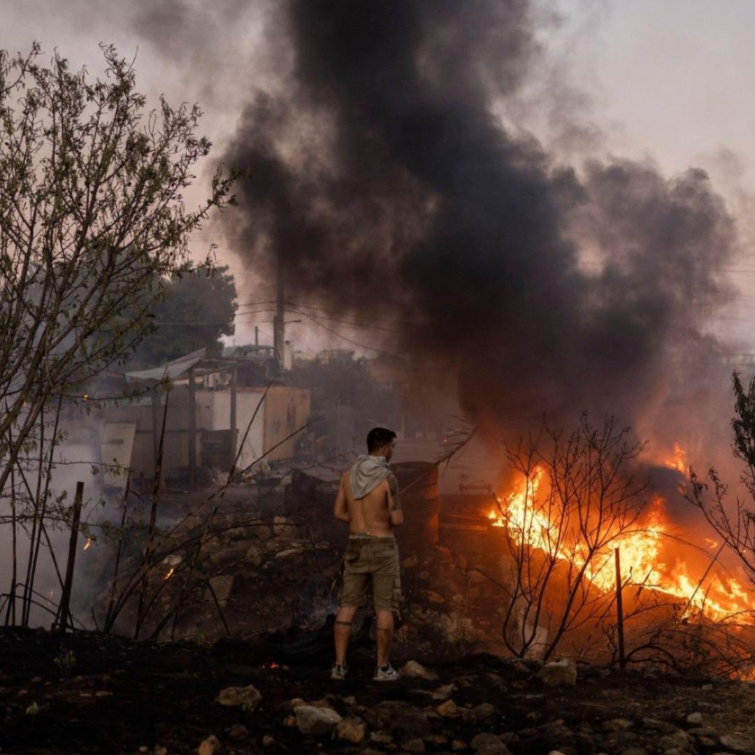 A man in a burned-out area looking at a home on fire.