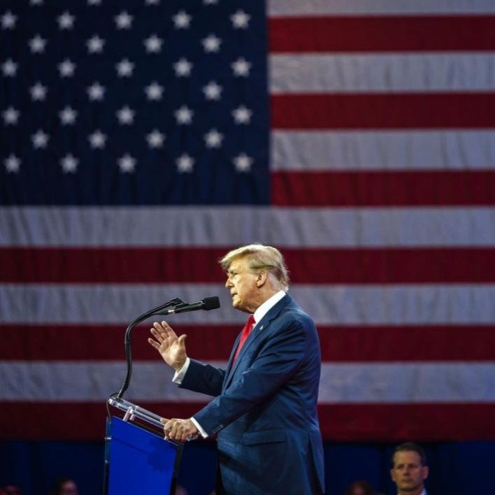 Donald Trump onstage at a podium in front of a large American flag.