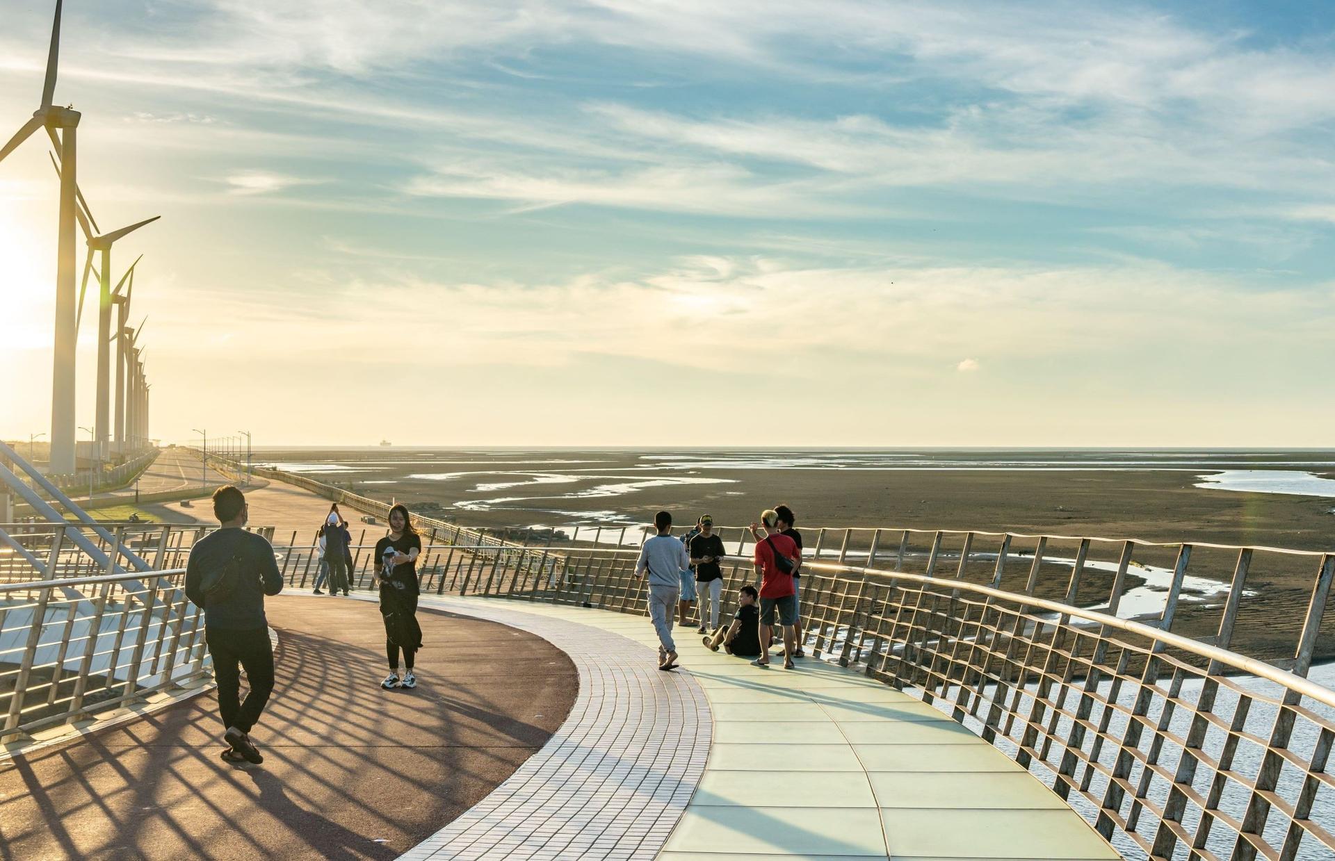 People walk on a bridge over wetlands with windmills in the distance